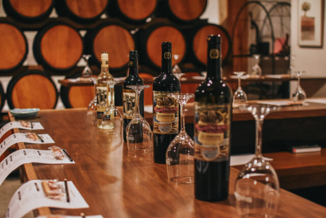 Bottles of JS Hamilton VQA from Pelee Island Winery line up with glassware on the tasting bar at the Pelee Island Winery Pavilion on Pelee Island with wine barrels in the background.